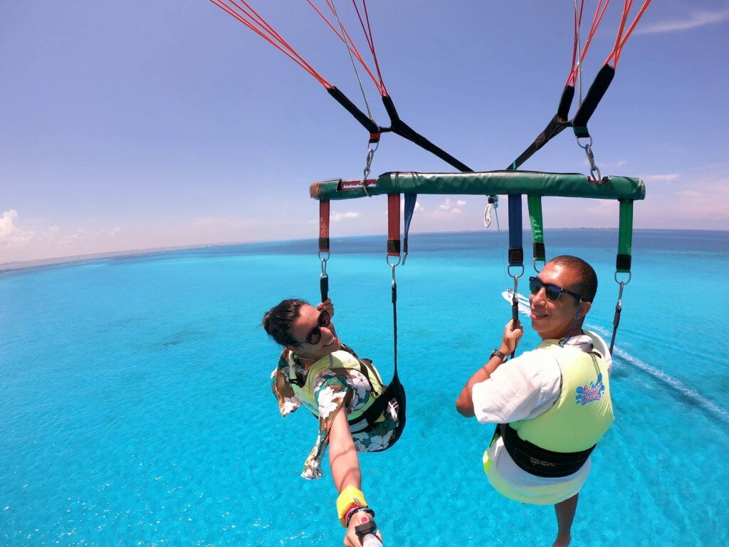 Foto de duas pessoas, uma mulher e um homem, suspensas em equipamentos de segurança em cima do mar azul. A mulher tem a pele clara e estende o seu braço para tirar a foto com um pau de selfie, ela usa óculos de sol e colete salva-vidas e o homem tem a pele escura, usa óculos de sol e também colete salva-vidas e está sorrindo para a foto. Ao fundo, vemos a água do mar num tom azul piscina, bem clara e limpa.